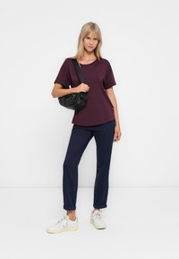 Ženska v temno vijolični majici, mornarsko modrih hlačah, belih supergah in črni torbi preko rame, stoji pred enobarvno belo ozadjem.