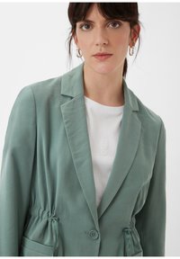 Lichtgroene blazer van soepele stof met een gestructureerde kraag, een getailleerde taille en twee voorkruiszakken. Draagt over een wit t-shirt.