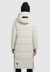 Hvit pufferjakke med hette og lange ermer, med horisontale quiltede seksjoner og sidelommer med glidelås. Inkluderer en svart logo-detalj på baksiden.