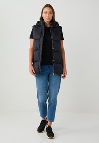 Gilet imbottito nero con cappuccio, zip dorata e bottoni a pressione, indossato sopra una maglia nera e abbinato a jeans blu e sneakers nere.