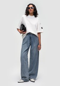 Camiseta blanca oversized, jeans de tiro alto de pierna ancha en azul claro, bolso negro y zapatillas blancas con detalles en negro. Gafas de sol oscuras completan el look.