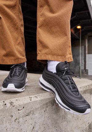 Person iført sorte og hvide Nike-sneakers med hvide sokker og brune bukser står på en betonrand udendørs.