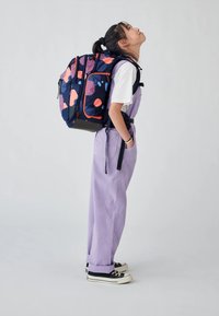 Rucksack mit blauer Basis, der pinke und blaue abstrakte Muster aufweist, mehrere Fächer hat und verstellbare Riemen bietet. Das Modell trägt lila Latzhosen.