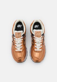 Zapatillas metálicas de bronce con cordones blancos, detalles en negro y acabado texturizado. Presentan un cuello acolchado y suela con amortiguación.