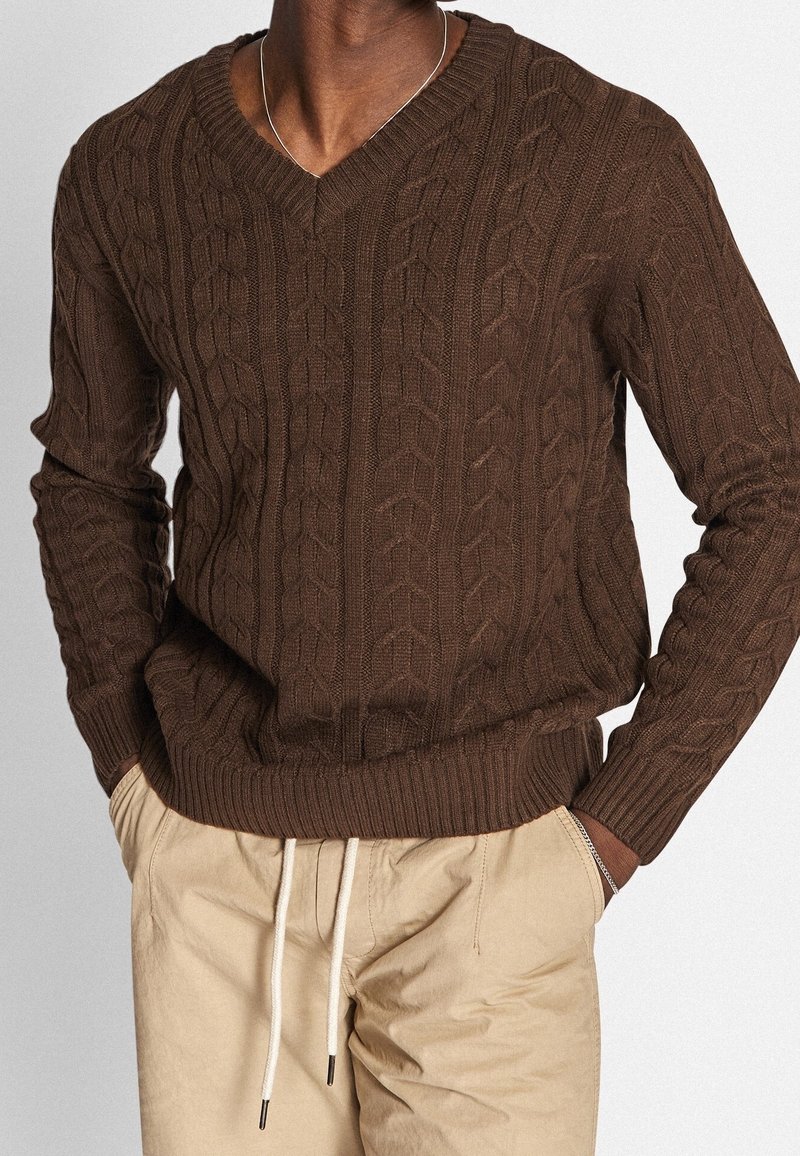 Pull marron tricoté à col en V style torsadé avec poignets et ourlet côtelés. Associé à un pantalon beige à coulisse, mettant en valeur le détail du tissu texturé.