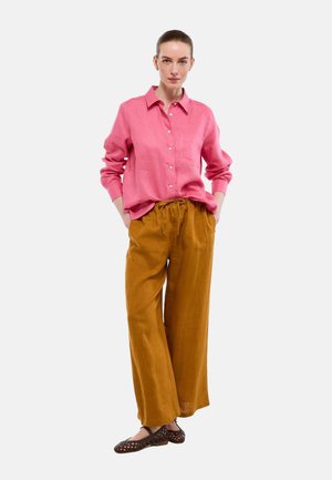 Femme debout, les mains dans les poches, portant une chemise ample rose à boutons, un pantalon large moutarde et des ballerines tressées marron sur un fond blanc.