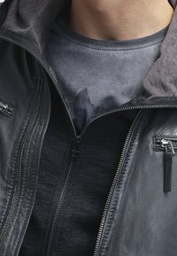 Zwarte leren jas met een grijze gewatteerde capuchon; beschikt over twee ritssluitingen en een gladde textuur, gedragen over een donker grafisch T-shirt.