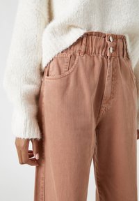 Bavlněné kalhoty s vysokým pasem, světle růžové, s dvěma předními kapsami, dvěma knoflíky a jemnou strukturou. Pleten�ý krémový svetr je částečně viditelný.