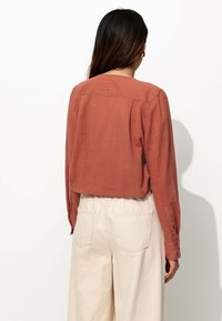 Blouse couleur rouille en tissu léger, avec une coupe décontractée, des manches longues et des poignets boutonnés, associée à un pantalon large crème.