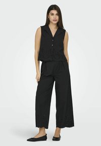 Top nero senza maniche con chiusura a bottoni e orlo a scallop; abbinato a pantaloni neri a gamba larga con dettagli in broderie anglaise.