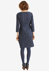 Marineblaues gepunktetes Kleid mit Dreiviertelärmeln, tailliertem Schnitt und knielangem Saum, ergänzt durch flache Knöchelschuhe und Strumpfhosen.