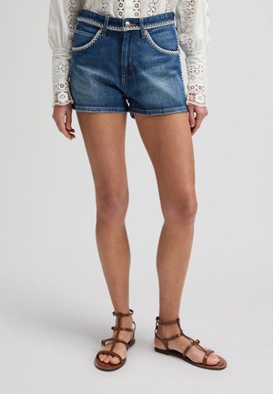 Kvinde iført blå denimshorts med hvid broderet kant, kombineret med en hvid bluse i hulmønster og brune flade sandaler med stropper.