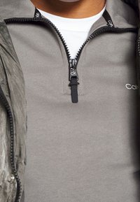 Grå zip-jacka i texturerat tyg, med en svart dragkedja med loggopull och en vit skjorta under.