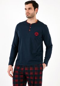 Camisa de manga larga azul marino con cuello de cuadros y botones rojos, que presenta un logo circular rojo en el pecho; combinada con pantalones de cuadros en rojo y negro.