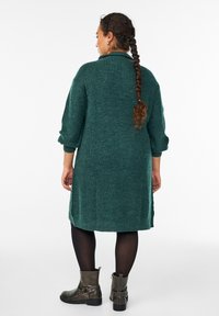 Žalias megztas kardiganas su aukštu apykakle, ilgomis rankovėmis ir šoninėmis skeltukais, derinamas su juodomis pėdkelnėmis ir pilkais kulkšnies batais.