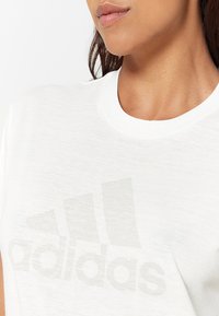 T-shirt bianca in cotone con il logo Adidas grigio chiaro stampato sul davanti. Il collo è di stile classico a girocollo, con una texture morbida.