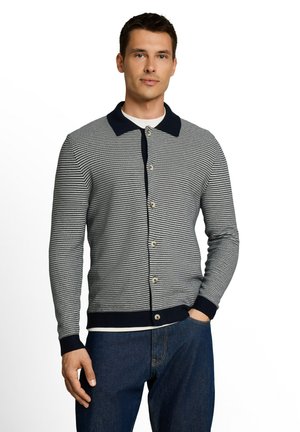 Mann trägt einen geknöpften, gestreiften Cardigan mit dunklem Kragen und Bündchen, weißem Unterhemd und blauen Jeans, steht mit einer Hand in der Tasche.