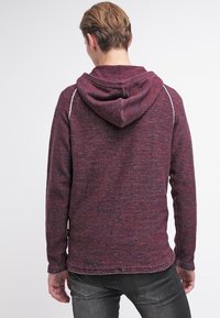 Mörkröd huvtröja i ett texturerat material, med synliga sömmar och en avslappnad passform. Huvan sitter bekvämt på bakhuvudet.