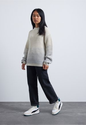 Jeune personne debout portant un pull en tricot crème, un jean noir et des baskets blanches, sur un fond gris clair uni.