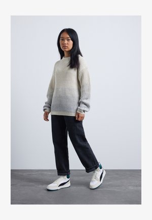 Jeune personne debout portant un pull en tricot crème, un jean noir et des baskets blanches, sur un fond gris clair uni.