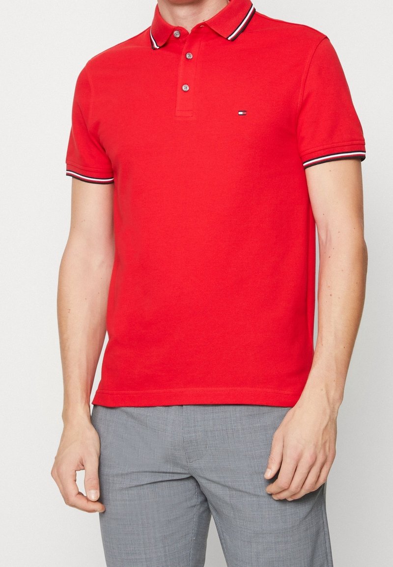 Homme portant un polo rouge avec un col et des manches rayés noir et blanc, associé à un pantalon gris à carreaux.