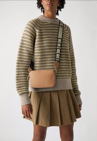 Tan krāsas ādas crossbody soma ar teksturētu virsmu, zelta aksesuāriem un rakstainu siksnu. Valkāta pār svītrainu, adītu svārku un plānu svārku.