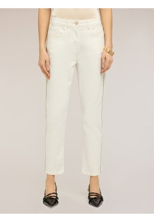 Kobieta nosząca białe jeansy o prostych nogawkach z bocznym łańcuszkiem, czarne szpilki z ostrym noskiem i złote bransoletki na prawym nadgarstku.