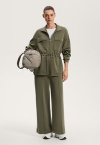 Olivno zelena zip-up jakna z dvema sprednjima žepoma, zategnjenim pasom in širokimi hlačami, ki se ujemajo. Bež torbica za ramo s podloženo zasnovo.