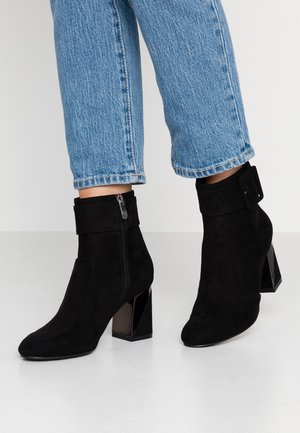 Bottes cheville en daim noir avec talons carrés et fermetures éclair latérales portées avec des jeans bleu clair retroussés sur un sol blanc.