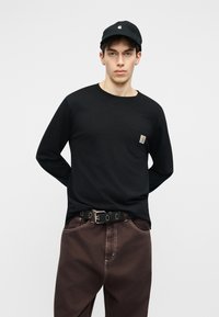 Νεαρός άνδρας που φοράει μαύρο μακρυμάνικο πουκάμισο με τσέπη στο στήθος, μαύρο καπέλο, καφέ παντελόνι με μαύρη ζώνη, στέκεται μπροστά σε απλό φωτεινό φόντο.