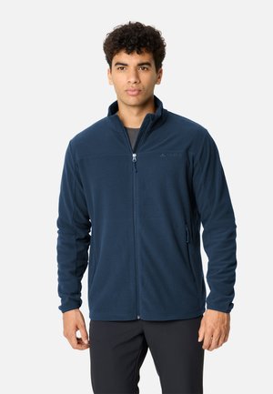 Jeune homme portant une veste polaire bleu marine zippée et un pantalon noir sur un fond clair uni.