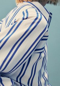 Persona che indossa una camicia bianca con grandi righe verticali e orizzontali blu, su uno sfondo azzurro chiaro.