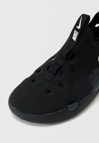 Svart slip-on-sko laget av fleksibelt materiale med perforert design, med en teksturert såle og en merket stropp på hælen.