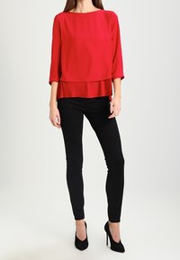 Blouse rouge à col rond et ourlet plissé, assortie à un pantalon noir slim et des escarpins noirs à talons hauts. Texture de tissu lisse.