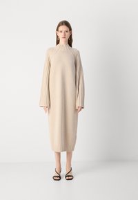 Beige kötött ruha bordás hosszú ujjal és magas nyakkivágással, egyenes fazonnal és térd alatti hosszúsággal. Fekete pántos szandál.