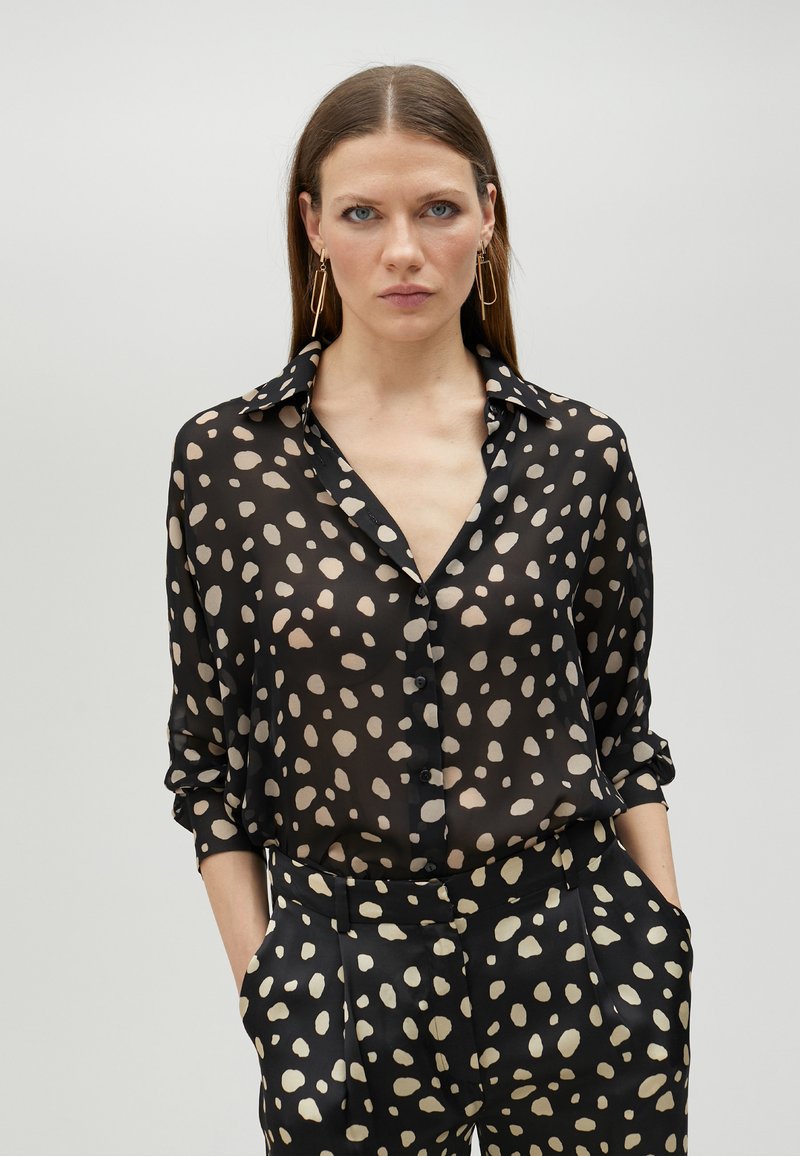 Sheer black blouse with beige polka dots, button-down front, and a classic collar. Features rolled-up sleeves and a loose fit.