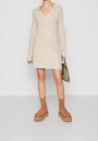 Beige stickad klänning med V-ringning och krage, långa ärmar och rak skärning. Matcher med bruna ankelstövlar och en mönstrad handväska.