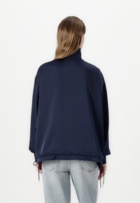 Navy modrá oversized bunda s vysokým límcem, texturovanou tkaninou, olemováním na šňůrku a dlouhými rukávy. Modelka má na sobě světle modré džíny.