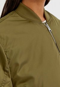 Close-up of a person wearing an olive green bomber jacket with a ribbed collar and a silver zipper partially zipped.