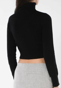 Maglione a collo alto a coste nere con maniche lunghe, design cropped, texture morbida, indossato con pantaloni grigi su misura dotati di tasca posteriore.