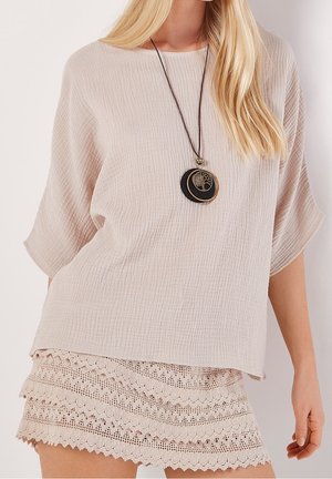 Femme portant un haut beige texturé à manches courtes, une jupe mini en dentelle superposée et un long collier pendentif avec un motif d'arbre.