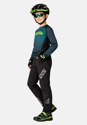 Kind mit grünem und schwarzem Fahrradhelm, Sonnenbrille, Handschuhen, langärmeligem Hemd, schwarzer Hose und grünen Fahrradschuhen, seitlich stehend.