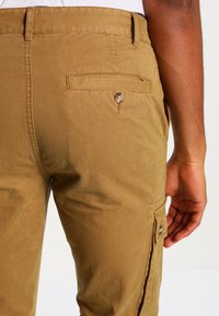 Close-up van een persoon die khaki broek draagt met een knoopzak aan de achterkant en een zijcargozak, hand ontspannen langs hun zij.