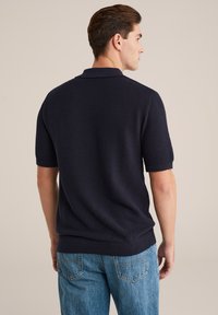 Vīrietis, ģērbies struktūrētā tumši zilos īsās piedurknes polo kreklā un zilos džinsos, stāv ar seju prom pret vienkāršu bēšu fonu.