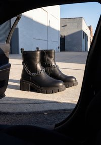 Paire de bottines en cuir noir avec talons épais et détail chaîne argentée posées sur un trottoir en béton près d'une porte de voiture.