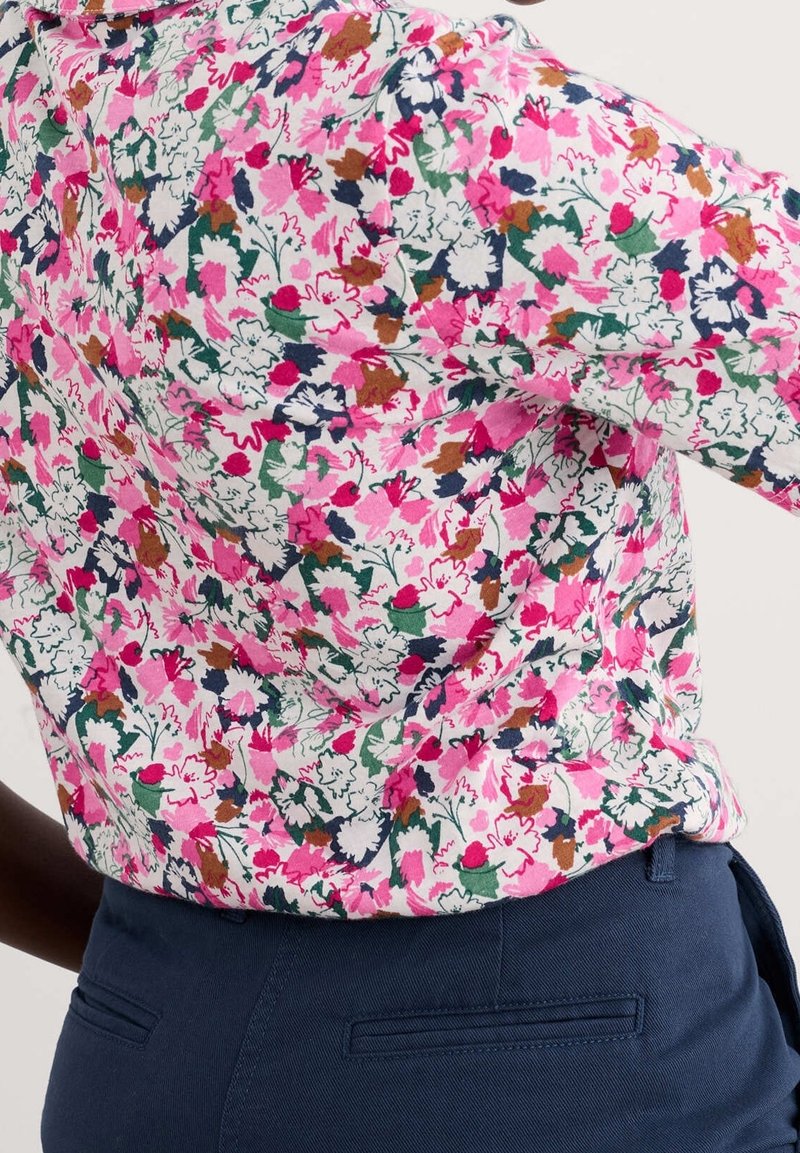 Personne portant une chemise à motifs floraux roses rentrée dans un pantalon bleu marine, montrant le dos et les détails de la taille.