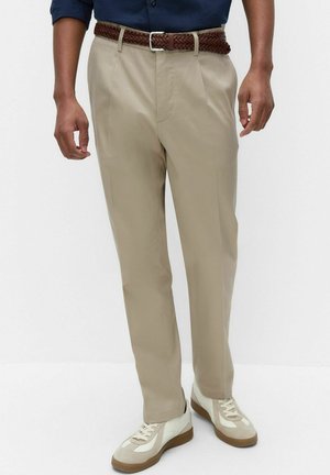 Mężczyzna w beżowych spodniach z fałdami z przodu, brązowym plecionym pasku, białych tenisówkach i granatowej koszuli z długim rękawem, wpuszczonej w spodnie.