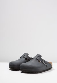 Sabots noirs avec une finition lisse et mate, dotés d'une sangle à boucle ajustable unique et d'une semelle en liège, reposant sur une semelle en caoutchouc texturé.
