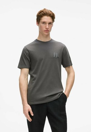 Jeune homme aux cheveux châtain clair portant un t-shirt gris foncé à manches courtes avec un petit logo, debout avec une main dans la poche.
