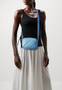 Borsa a tracolla azzurro chiaro in materiale liscio, con motivo a quadrati obliqui, chiusura a zip e tracolla regolabile. Ideale per un uso casual.